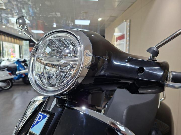 Close-up of the front of a black scooter, featuring a chrome headlight and controls, with blurred scooters visible in the background inside a dealership.