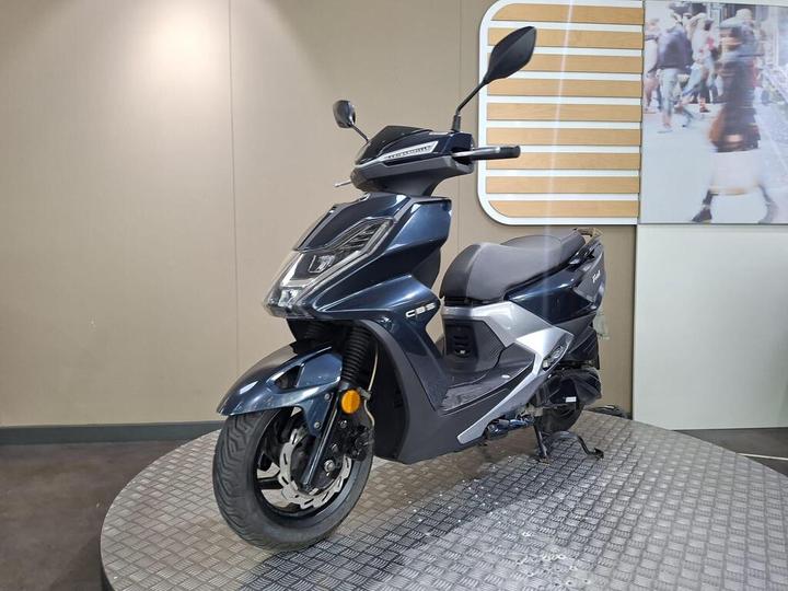 A dark blue modern scooter displayed on a rotating platform against a neutral wall, highlighting its sleek design and features.