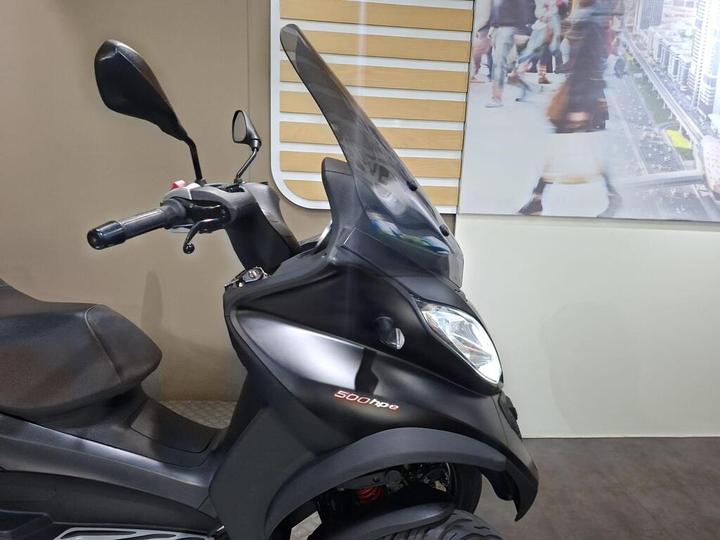 A close-up view of a black scooter featuring a prominent windshield, handlebars, and a smooth seat, set against a backdrop of a blurred cityscape with people walking.