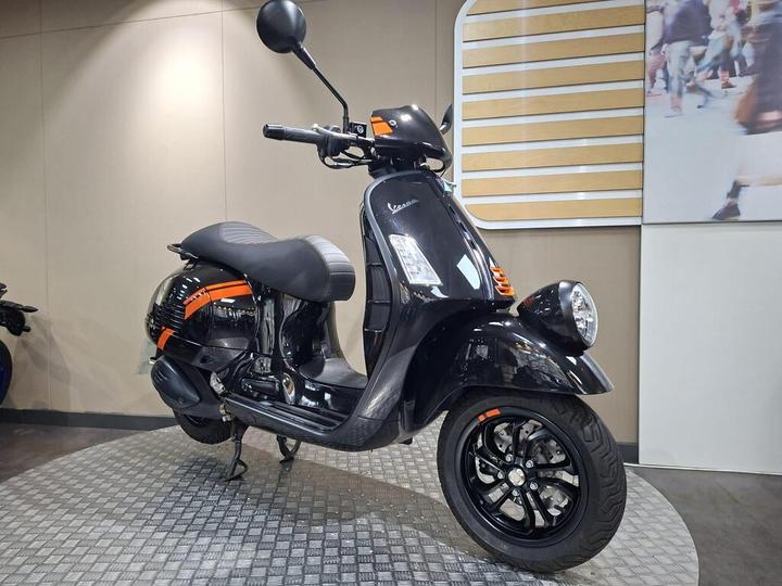 A sleek black scooter with orange accents stands on a textured gray surface, displayed indoors against a neutral background with decorative elements.