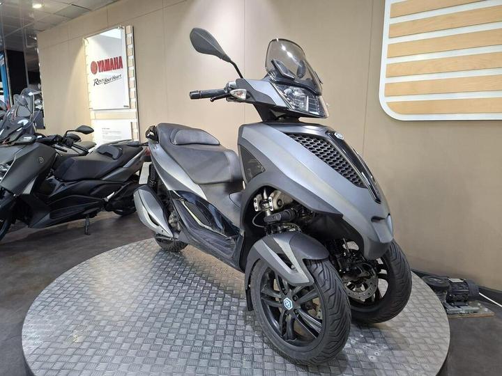 A sleek gray three-wheeled scooter is displayed on a textured platform, with a modern interior setting in the background featuring Yamaha branding and signage.