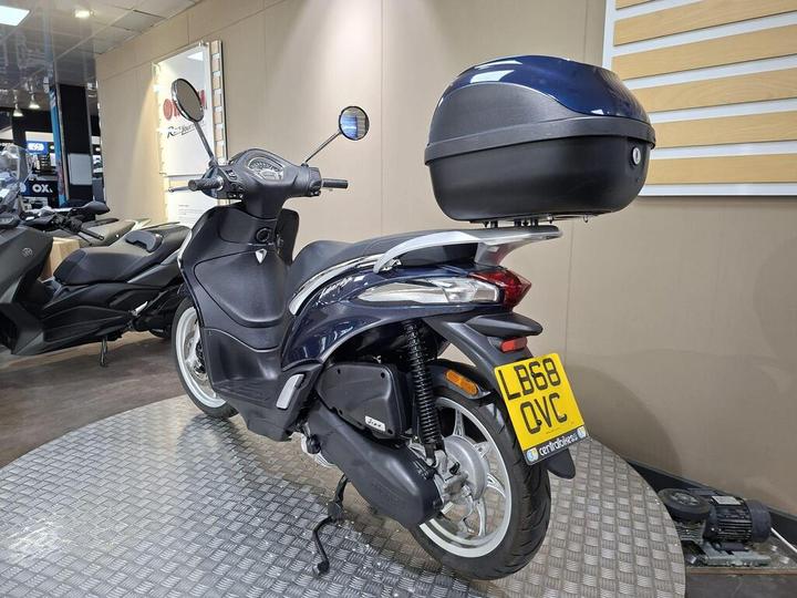 A blue and silver scooter is parked on a metal platform inside a showroom, featuring a top case. The back license plate reads "LB68 OVC." Other scooters are visible in the background.