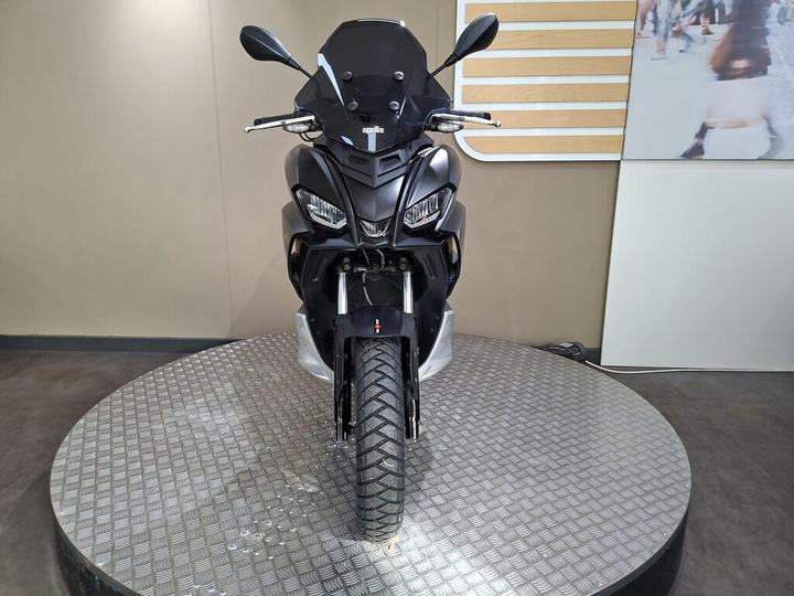 A sleek black scooter displayed on a rotating stand, featuring a prominent front view with modern design elements, a tinted windshield, and a textured tire.
