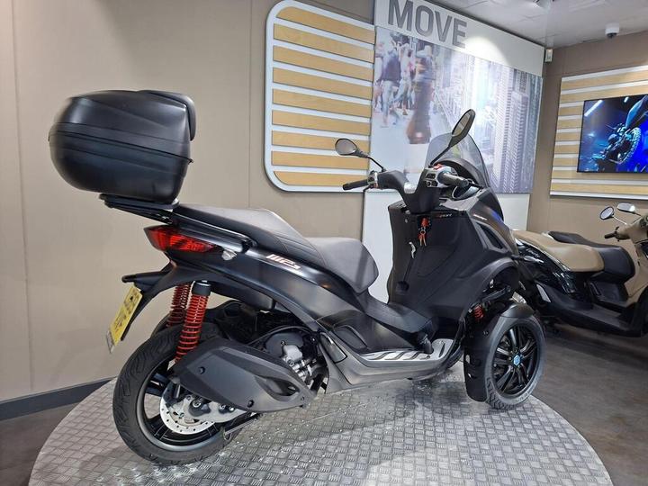 A black scooter with a rear storage box, parked on a metallic checkered surface in a modern showroom displaying various motorcycles and a large promotional image on the wall.