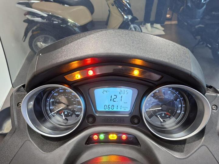 Dashboard of a scooter displaying speedometer and tachometer readings, with indicators showing battery voltage (121 volts) and total mileage (6011 miles). The temperature gauge and warning lights are also visible.