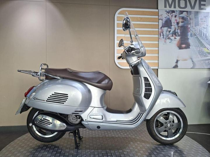 The Vespa GTS 300 Super 2016 A silver Vespa scooter with a brown seat is displayed against a neutral background. The scooter features a classic design with a chrome rearview mirror and distinctive body lines.