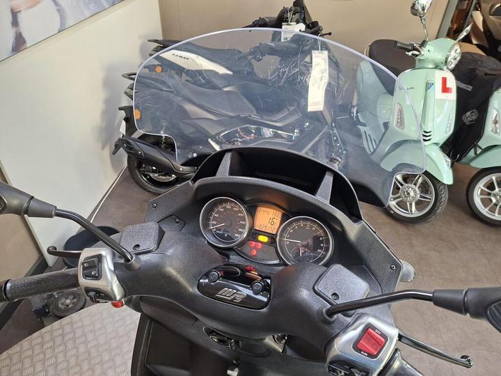 A close-up view of a scooter's dashboard, featuring a windshield, speedometer, and control levers, with additional scooters visible in the background.