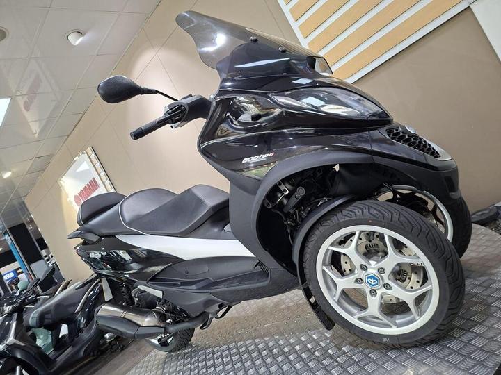A black and silver three-wheeled scooter is displayed in a dealership, showcasing its sleek design and modern features against a neutral background with showroom elements.