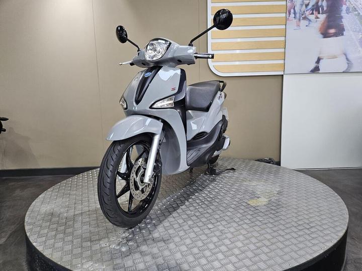 A gray scooter is displayed on a circular metal platform in a showroom setting, featuring a sleek design and modern aesthetics.