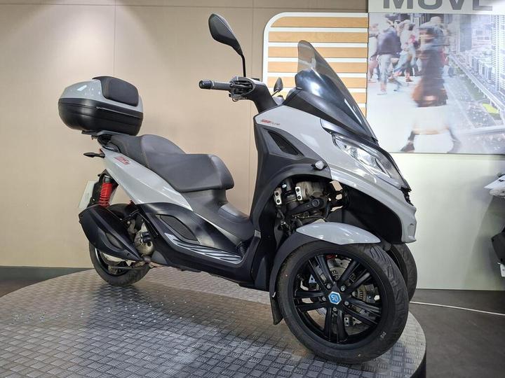 A gray three-wheeled scooter is displayed on a circular platform against a neutral-colored wall, featuring a storage case on the rear and a sleek design with a prominent front windshield.