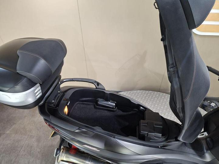 A close-up view of a scooter's seat and storage compartment, featuring a sleek design with a dark color scheme and visible interior lighting. The seat is partially reclined, showing the spacious storage area beneath it.