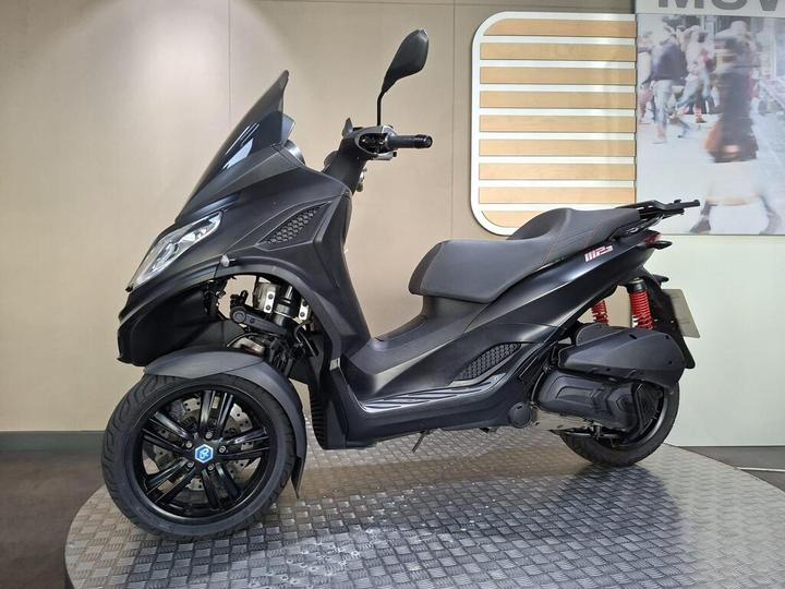 A black three-wheeled scooter is showcased on a textured surface, featuring a streamlined design, modern headlights, and red suspension accents. The background includes a blurred image of pedestrians.