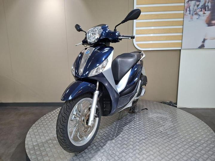 A blue scooter is parked on a circular, textured platform in a spacious showroom, showcasing its sleek design and chrome accents. The background features a neutral wall with a decorative panel.