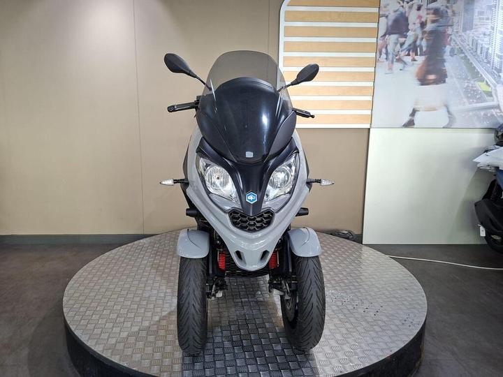 A grey and black three-wheeled scooter is displayed on a rotating platform against a neutral background, showcasing its front view with a visible windscreen and headlights.