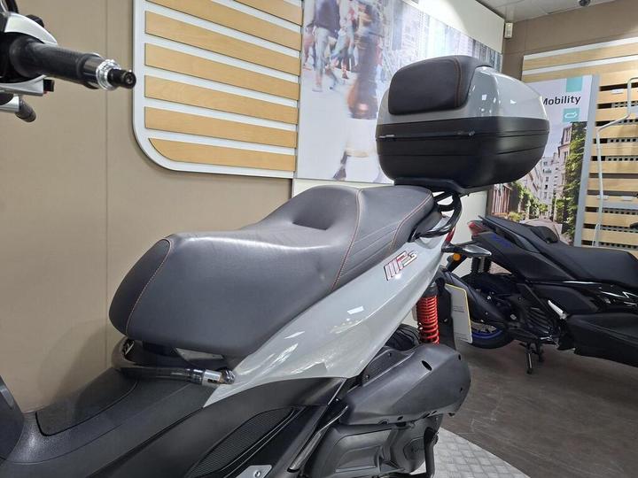 A close-up view of a modern scooter featuring a grey seat with red stitching and a rear storage box, displayed in a showroom with a light-colored wall and additional scooters in the background.