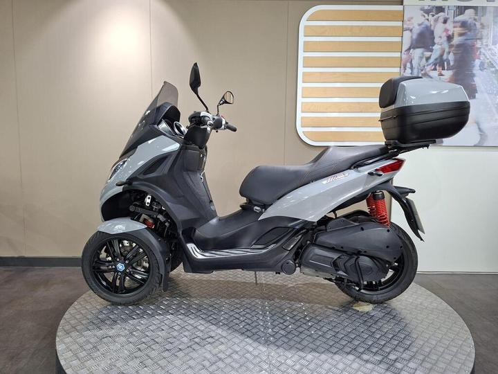 A gray three-wheeled scooter displayed on a circular platform, featuring a sleek design, protective windshield, and a rear storage compartment. The background includes a neutral-colored wall.