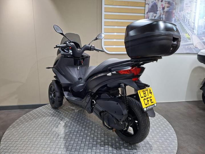 A black three-wheeled scooter is displayed in a showroom, featuring a rear storage box and a license plate. The background includes a light-colored wall and an abstract cityscape.