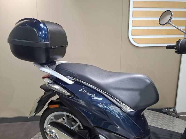 A close-up side view of a blue scooter featuring a comfortable black seat and a rear storage box, set against a neutral background with horizontal wooden slats.