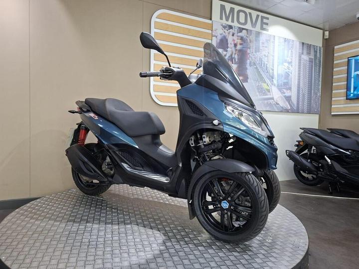 A blue three-wheeled scooter is displayed on a circular stand with a textured surface, set against a light beige wall featuring a "MOVE" sign and a backdrop of a busy cityscape.