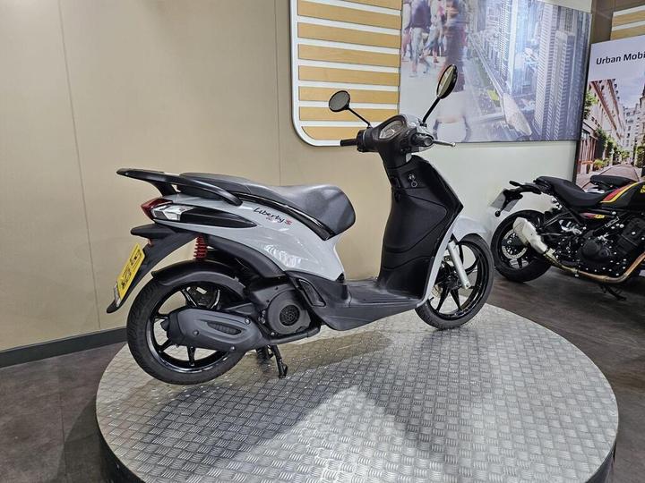 A sleek gray and black scooter displayed on a round, metallic platform in an exhibition space, with a blurred backdrop featuring urban imagery and another motorcycle on the right.
