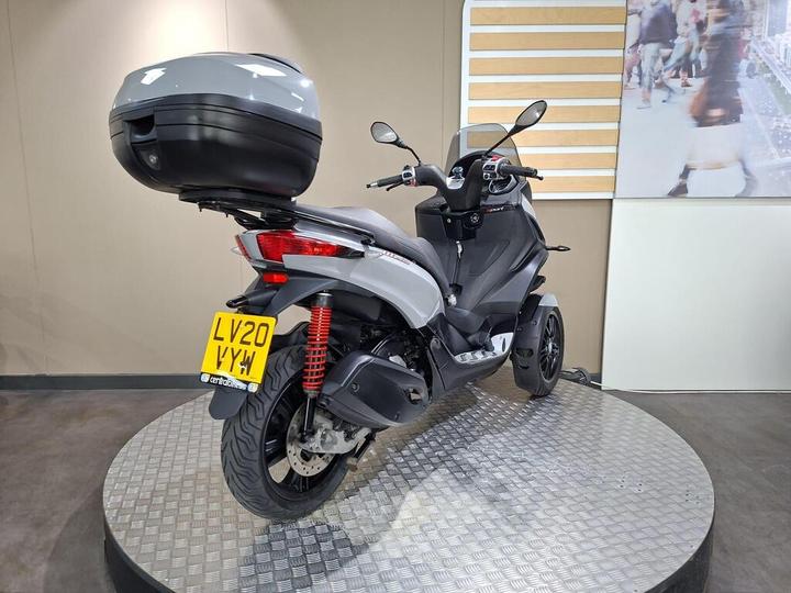 A gray scooter with a black and silver top case, displaying the license plate LV20 VYW, is positioned on a metallic circular platform in a showroom setting.