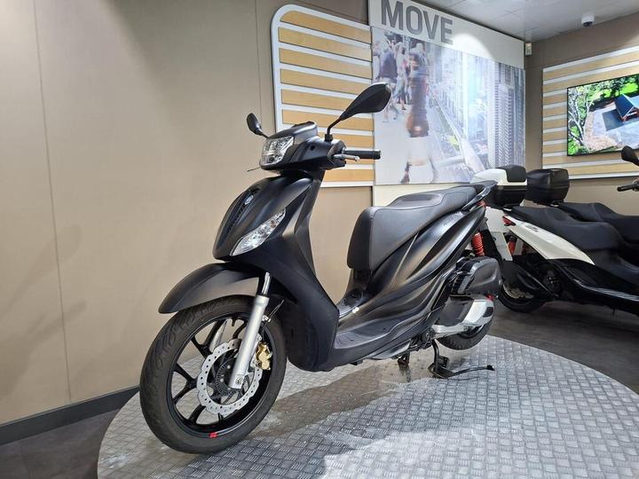 A black scooter displayed on a checkered floor, with a modern interior featuring a wall banner that says "MOVE" and an LCD screen showing green outdoor images in the background.