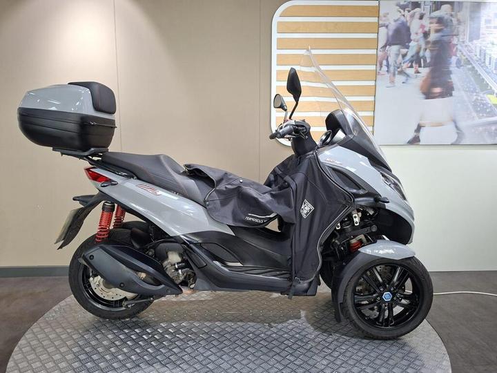 A gray three-wheeled scooter with a black storage box on the rear, partly covered with a protective cover, is displayed on a circular metal platform against a light-colored wall with a large image of a busy street scene in the background.