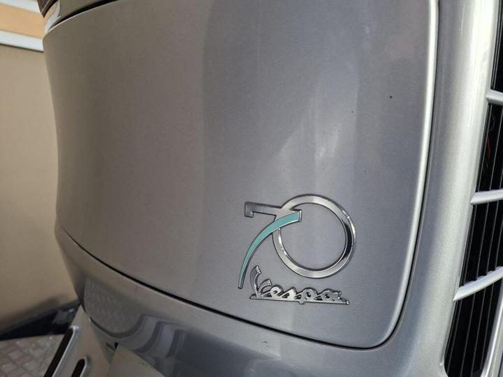 Close-up of a silver Vespa scooter logo featuring the number "70" in a circular design, celebrating its 70th anniversary.