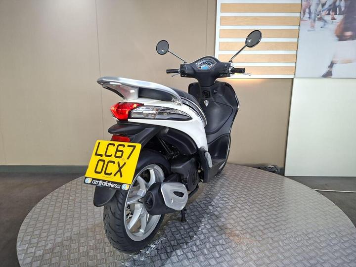 A silver and black scooter with a visible rear view, showcasing the license plate "LC67 OCX," parked on a textured circular platform in a modern interior setting.