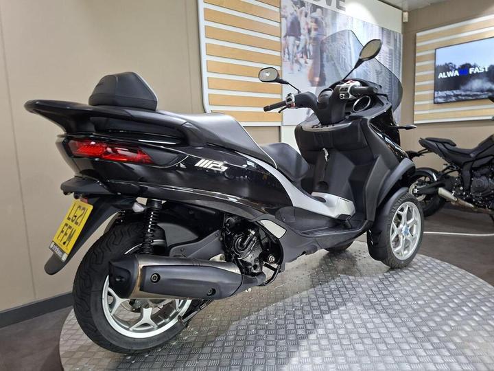 A sleek black three-wheeled scooter displayed in a showroom, featuring modern design elements and a rear view showcasing its rear tire, exhaust, and seat. The background includes a light-colored wall with promotional graphics.