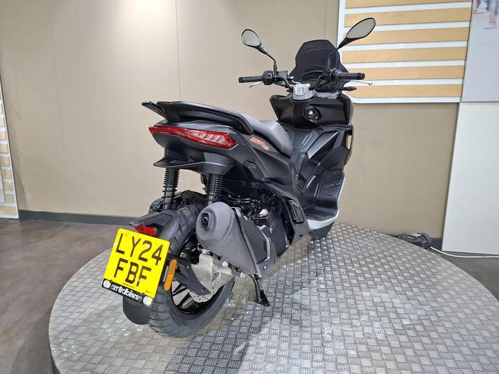 A sleek black scooter with a modern design, featuring a prominent rear light, mounted on a circular display platform in a showroom setting. The license plate reads "LY24 FBF."