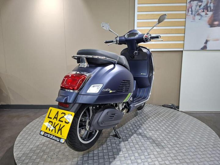 A blue scooter is displayed on a circular platform, showcasing its rear profile. The license plate is visible, reading "LA25 RKK." The background features a neutral wall with light wood accents.