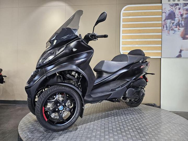 A sleek black scooter with a large windshield and distinct wheel design is displayed on a rounded platform against a light-colored wall.