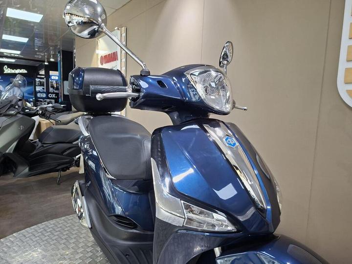 A close-up view of a blue scooter featuring a sleek design, prominent headlight, and rearview mirror, displayed in a modern showroom.