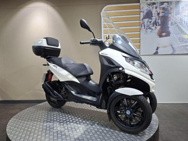 A white three-wheeled scooter with a clear windshield and rear storage compartment is displayed on a circular metal platform against a neutral wall, featuring a blurred background of people walking.