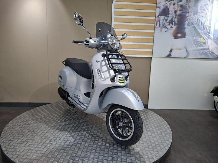 A silver scooter displayed on a circular, textured platform, featuring a transparent windscreen, dual mirrors, and a prominent front headlight. The backdrop showcases a vibrant urban scene.