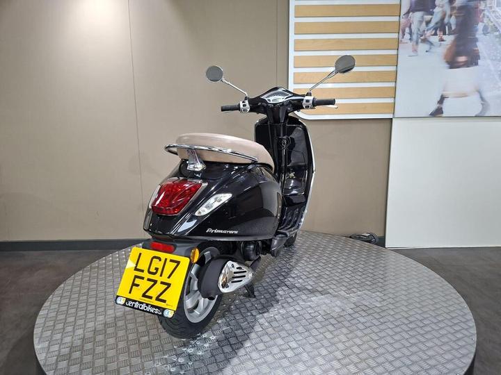 A black scooter with a beige seat is displayed at an angle, showcasing the rear side and license plate, set against a neutral background.