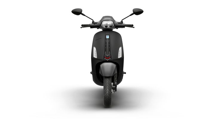 A sleek, black scooter is shown from the front, featuring a minimalist design, modern headlights, and a prominent logo. The scooter's front wheel is visible, resting on a gray surface with a subtle shadow.