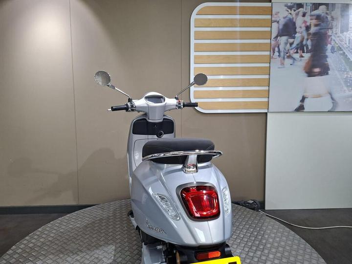 A silver scooter displayed in a showroom, viewed from the rear. The scooter features a sleek design with side mirrors and a rounded tail light. A light-colored wall with a wooden panel accent is visible in the background, alongside a blurred image of pedestrians.