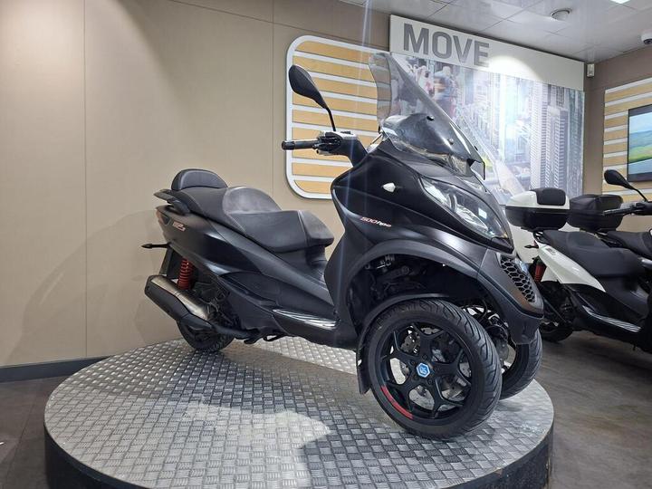 A black three-wheeled scooter displayed on a circular platform in a showroom, featuring a windshield, comfortable seating, and distinctive wheel design. The background includes branding elements and a cityscape image.