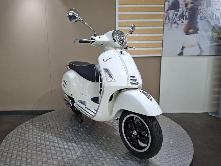 A stylish white Vespa scooter displayed on a circular platform against a neutral background. The scooter features chrome mirrors, a retro design, and black accents on the seat and wheels.