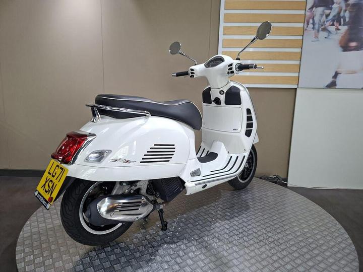 A white scooter with a black seat, displayed on a round metallic platform, set against a neutral background. The scooter features a retro design with chrome accents and a visible license plate.