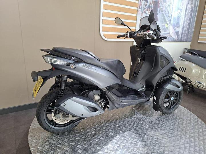 A grey scooter with a distinct design, featuring a prominent front body and a single front wheel, is displayed on a circular metallic platform. The background includes wooden panels and promotional images of urban scenes.