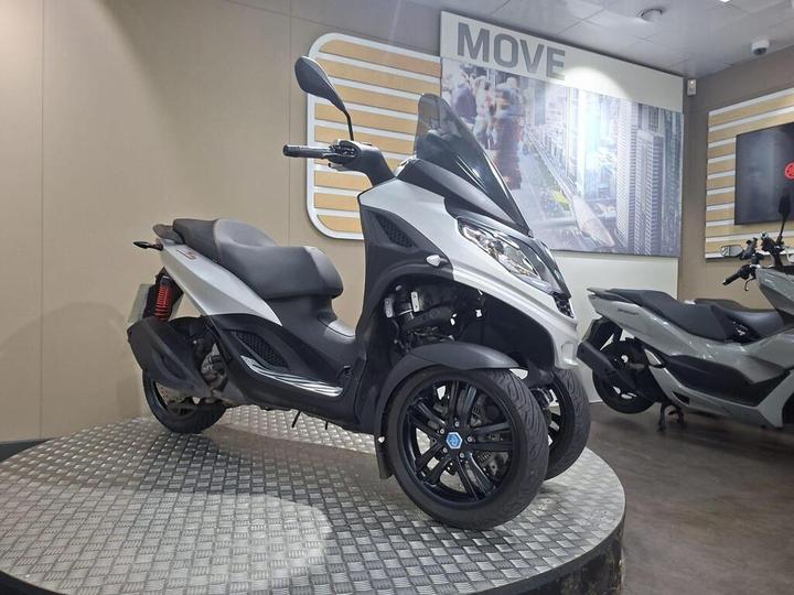 A silver three-wheeled scooter displayed on a circular metal platform in a showroom, with a background featuring a cityscape and the word "MOVE."