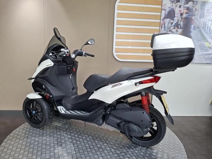 A white and black three-wheeled scooter is showcased in a modern display environment, featuring a streamlined design, a rear storage box, and detailed red suspension. The background includes a blurred image of a busy urban scene.