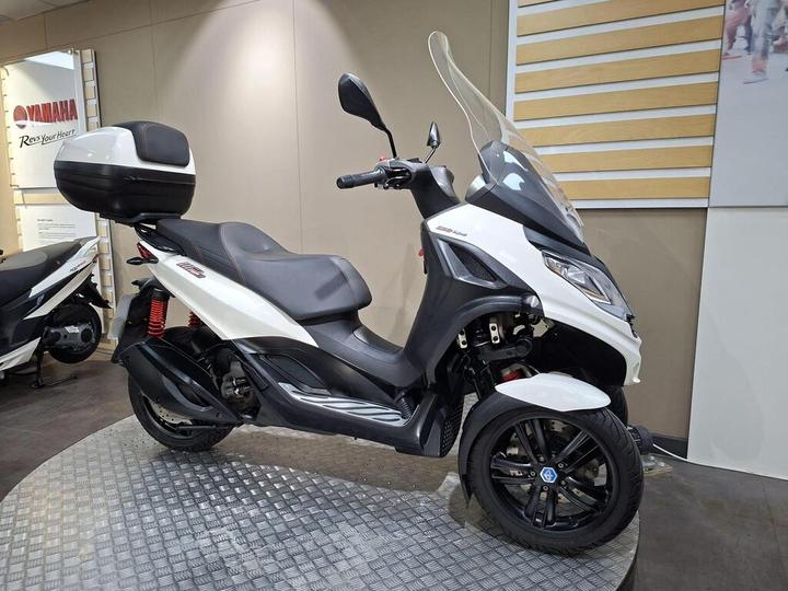 A white and black three-wheeled scooter with a tall windshield, side mirror, and a rear storage case, displayed in a showroom with a modern interior and Yamaha branding in the background.