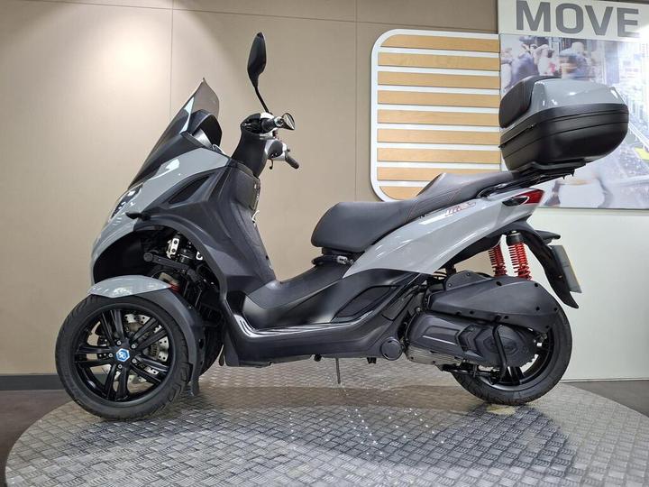 A gray three-wheeled scooter displayed on a textured platform in a modern showroom, with a windshield and storage compartment on the rear.