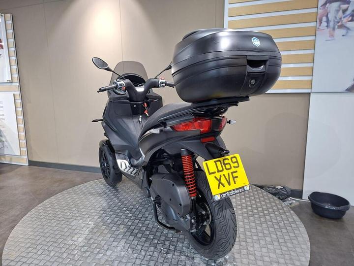 A black scooter with a top case is displayed in a showroom, showcasing its rear view and license plate reading "LD69 XVF." The background features neutral-colored walls and flooring.