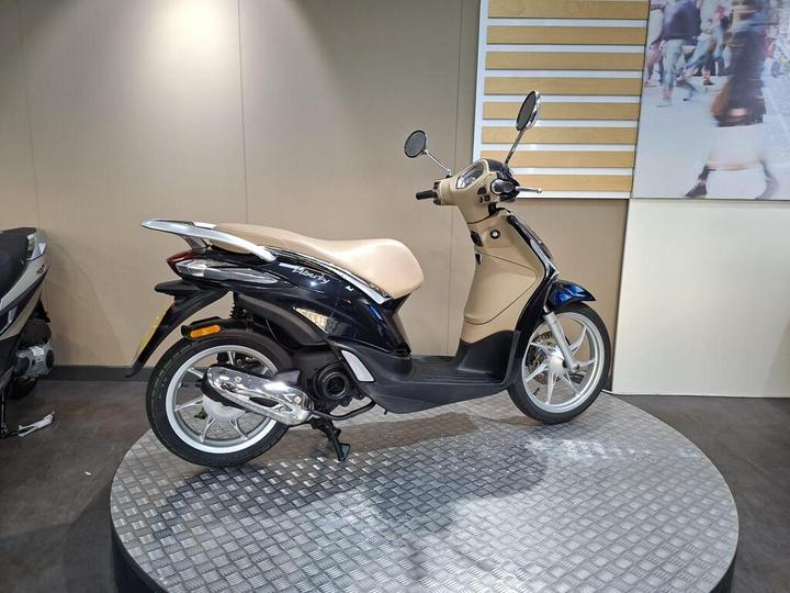 A stylish black and beige scooter displayed on a circular platform in a showroom, with a soft, minimalist background.