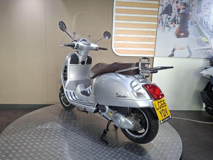 A silver Vespa scooter positioned on a circular platform in a showroom, featuring a brown seat and a clear windshield, with a subtle urban backdrop visible in the background.